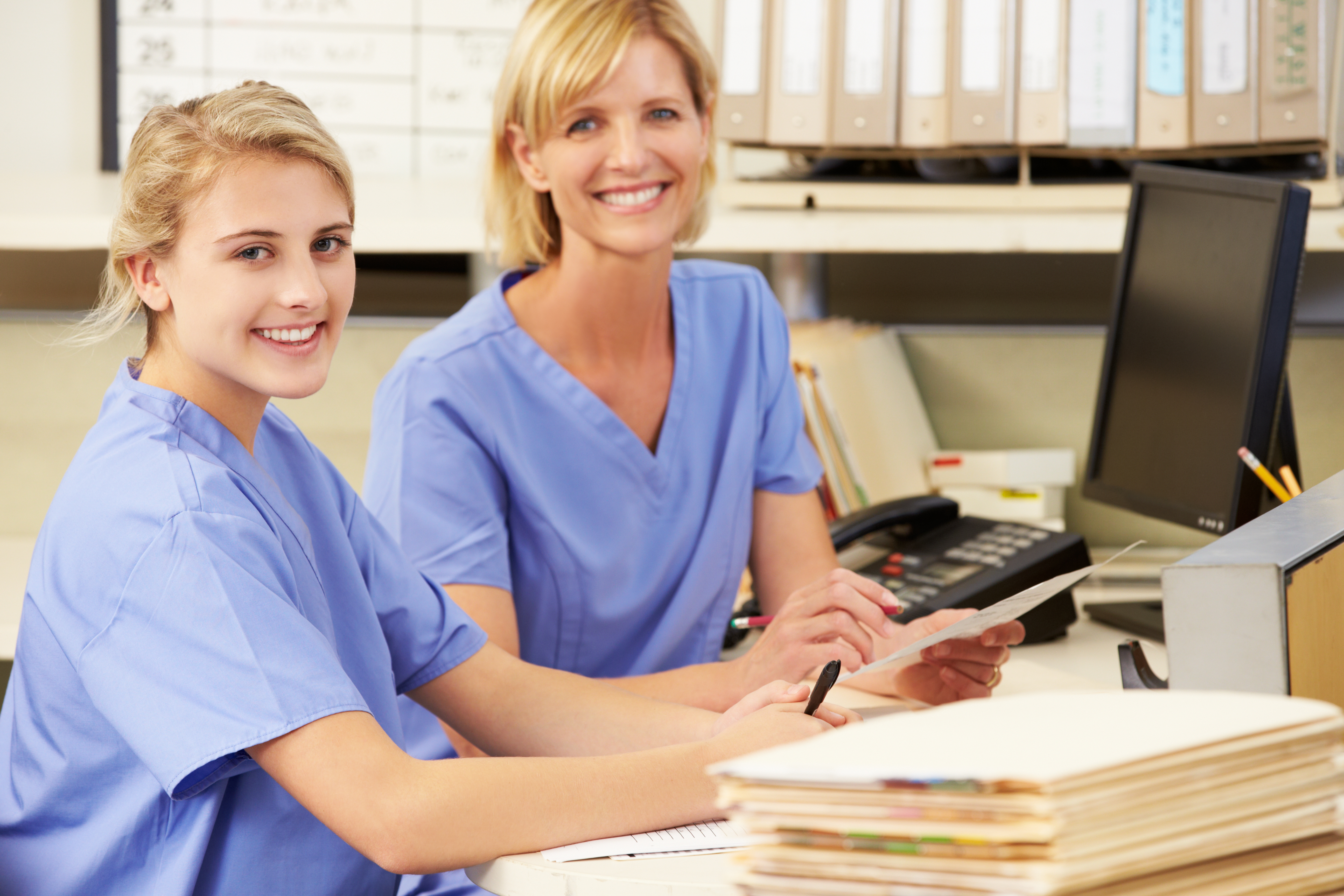 Zwei Pflegehelferinnen sitzend lächeln am Schreibtisch. 